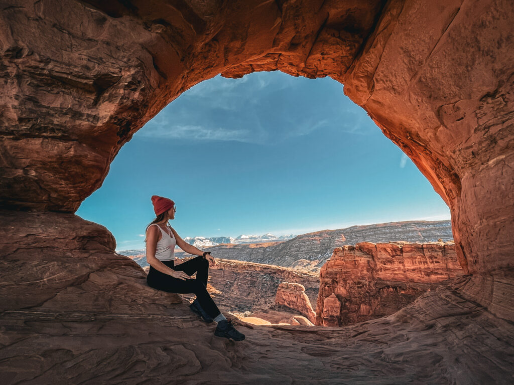 Kate, the owner of Kate's Crossing Blog, sits in a rocky archway, looking out over a breathtaking landscape of red rock formations and distant snow-capped mountains.