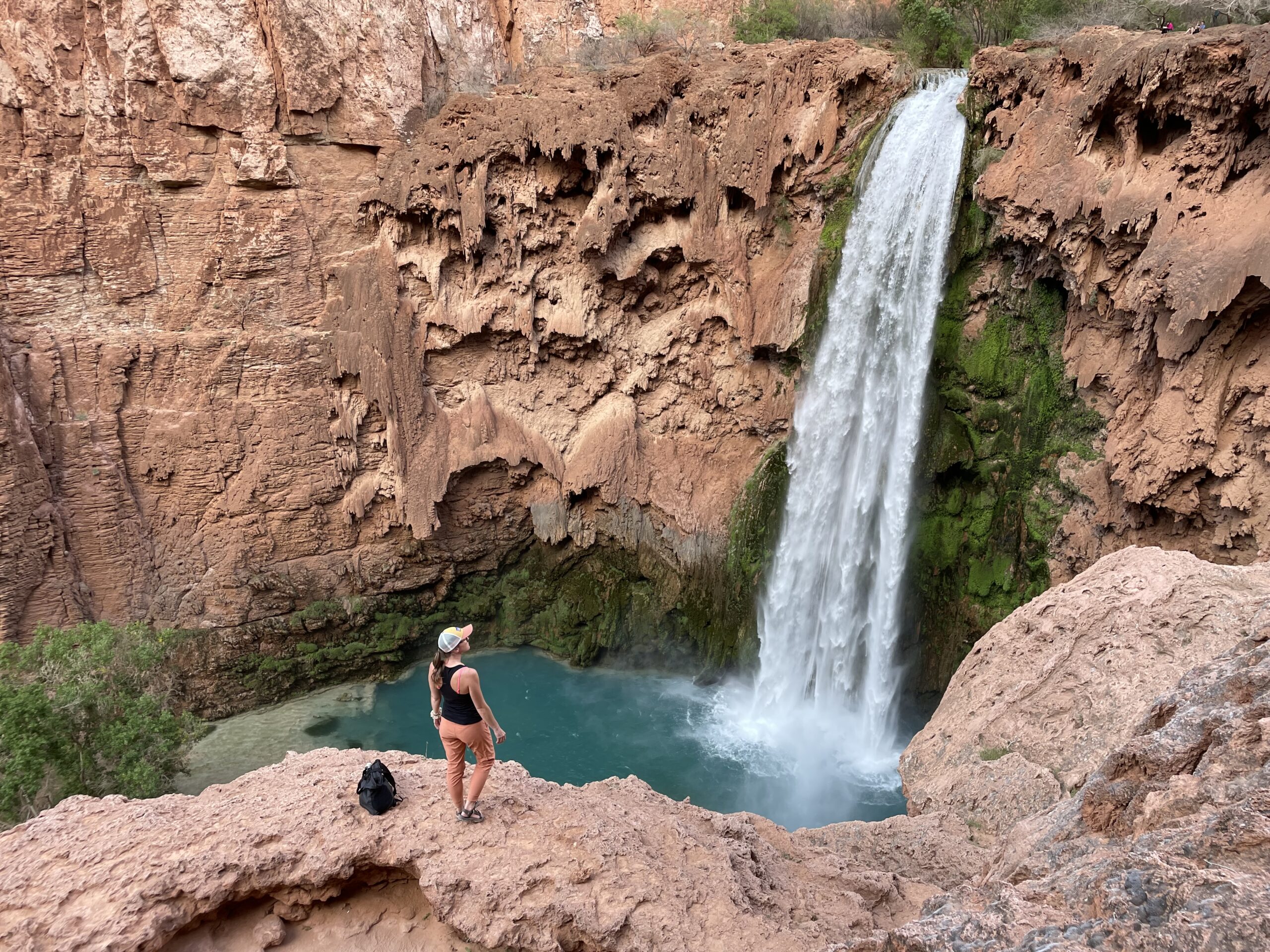 Kate standing on a cliff edge, gazing at the stunning turquoise waters of Mooney Falls in Havasu, Arizona, surrounded by red rock and green moss.