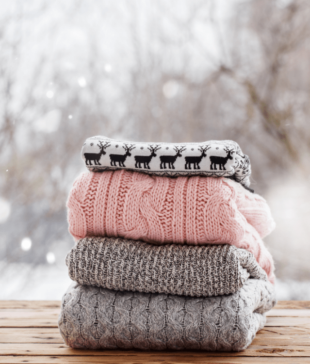 A stack of cozy winter sweaters in various shades of gray and pink, topped with a festive reindeer pattern, set against a snowy, blurred background.