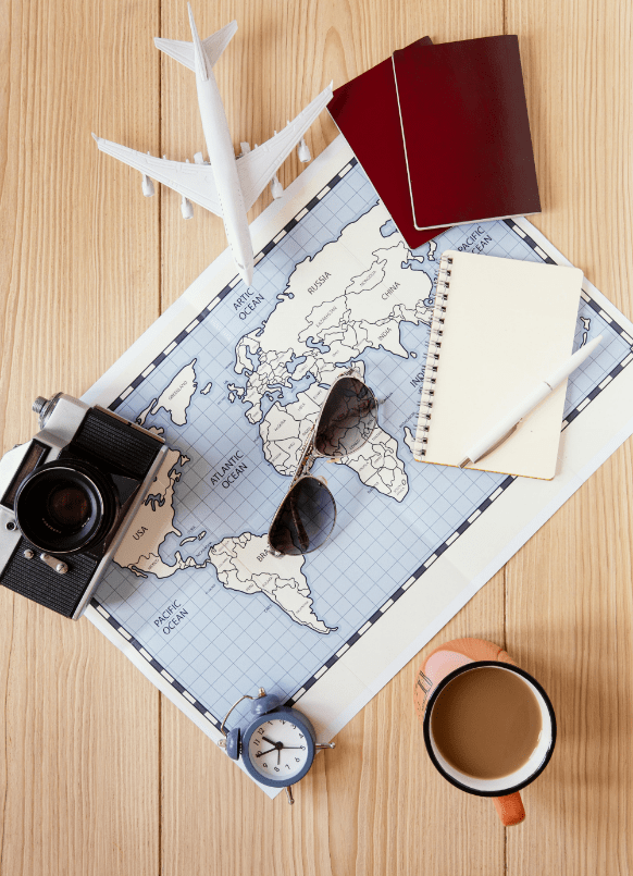 A travel flat lay featuring a world map, sunglasses, a camera, a model airplane, passports, a notebook with a pen, an alarm clock, and a cup of coffee on a wooden surface.