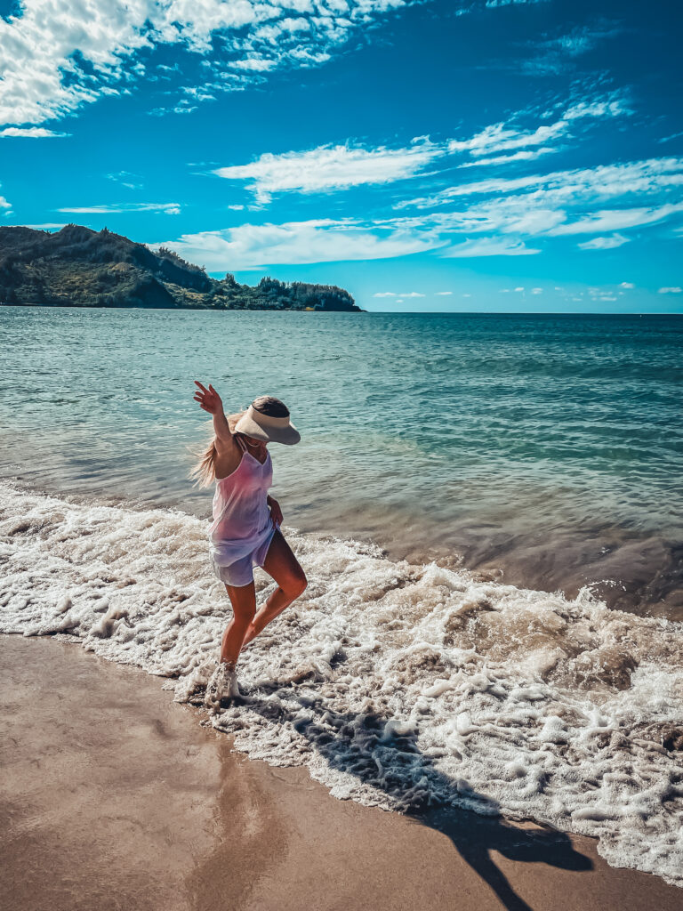 Kate playing in the waves on a sandy beach in Kauai, Hawaii, with a clear blue sky and lush green hills in the background.