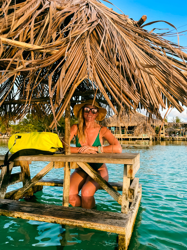 Kate sitting under a thatched roof cabana out in the water of Belize, wearing a green swimsuit and a sun hat, with a dry bag keeping her belongings safe.