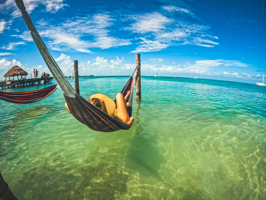 Kate relaxing in a hammock over clear turquoise water, wearing a sun hat, with a pier and boats in the background, showcasing a perfect beach day in Belize.