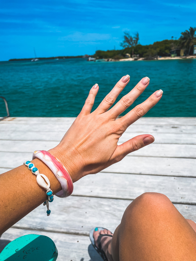 Close-up of Kate's hand wearing a pink conch shell bracelet, with a beautiful ocean view in the background, highlighting a unique accessory to include in your Belize packing list.