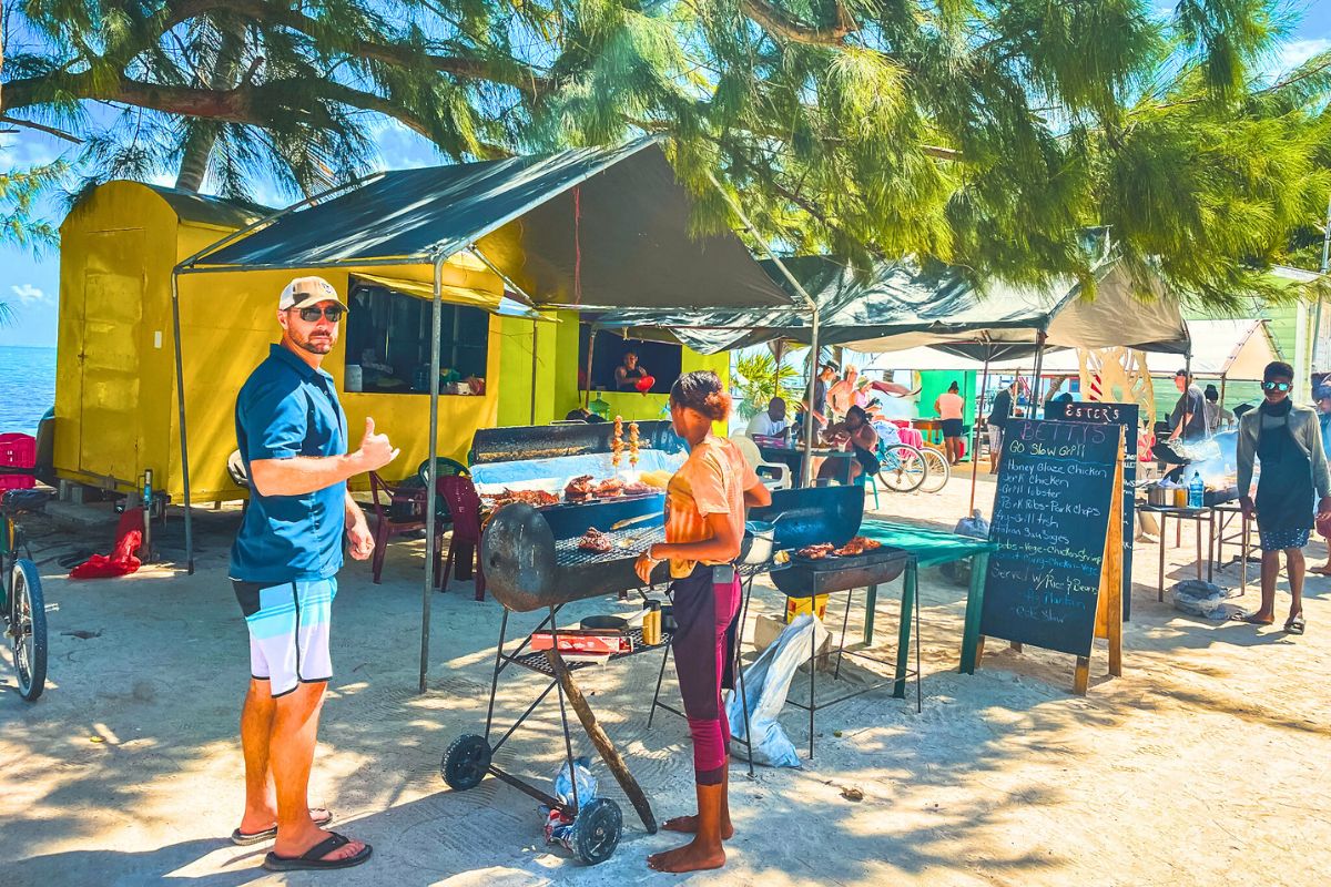 Kate's husband in a blue shirt and shorts gives a thumbs-up next to a woman grilling food under a canopy at a beachside food stall. Shows a belize travel tip about making purchases. The stall has a yellow building and a chalkboard menu listing items like honey glaze chicken, jerk chicken, and grilled lobster. People are dining and socializing under the shade of trees, with the sea visible in the background.