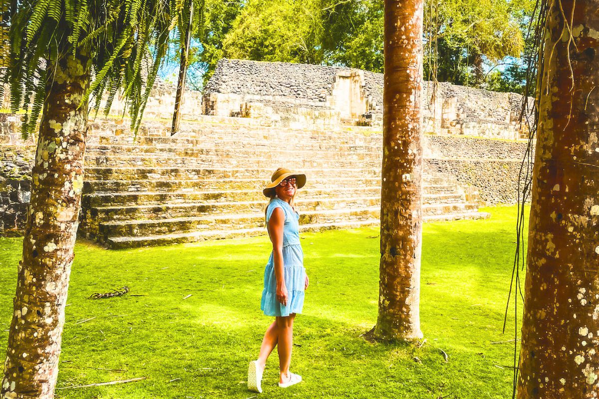 Kate in a blue dress and wide-brimmed hat stands between trees, smiling, with ancient stone ruins and lush green grass in the background.