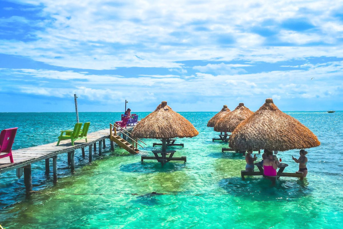 Thatched-roof palapas stand over clear turquoise water with people seated around submerged tables. A wooden dock with colorful chairs extends into the sea, where a person sits enjoying the view under a partly cloudy sky.