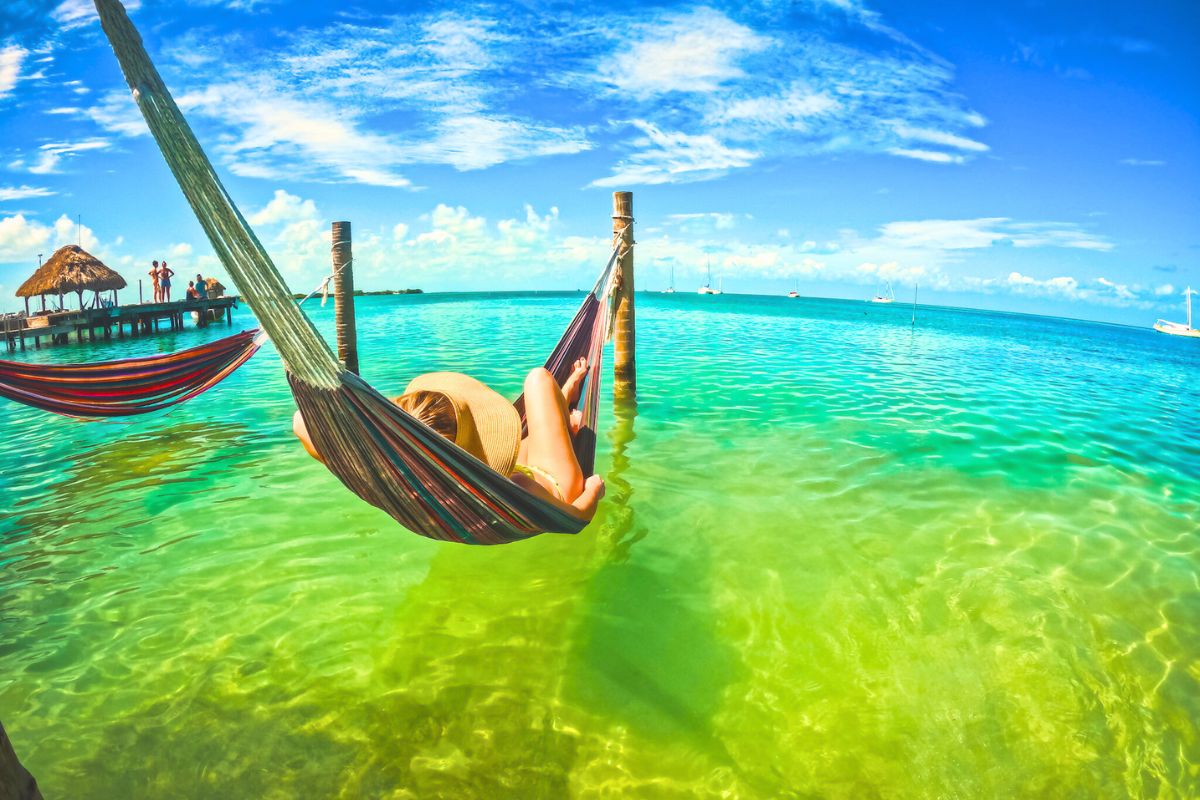 Kate in a wide-brimmed hat relaxes in a hammock suspended over clear turquoise water of Caye Caulker Belize. The scene features a sunny sky with fluffy clouds, a distant dock with thatched-roof palapas, and boats anchored further out in the sea.