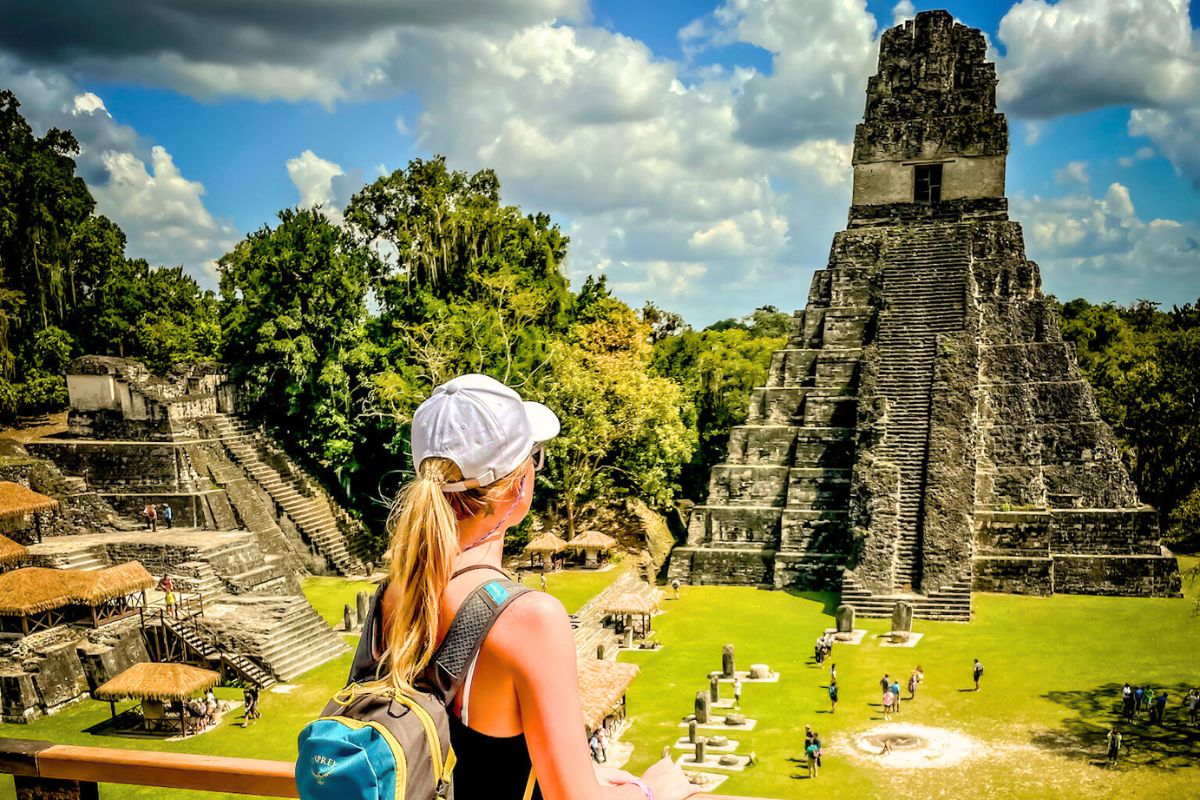 Kate with a backpack and a white cap gazes at a large ancient Tikal pyramid and surrounding ruins under a partly cloudy sky. The scene is lush with greenery and features several smaller structures, with tourists exploring the historical site.