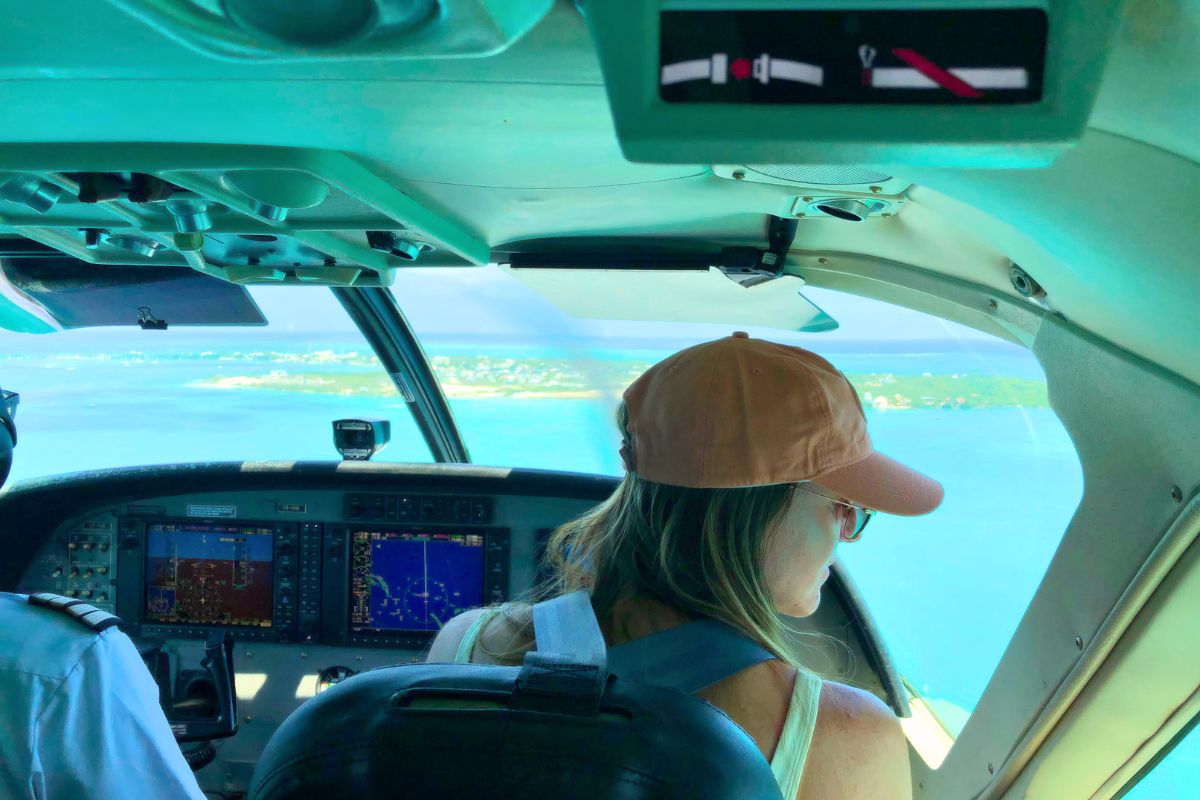 Kate in a baseball cap looks out the window from the co-pilot seat of a small airplane. The pilot is visible on the left, and the cockpit displays show flight information. Below, Belize's clear turquoise waters and small islands are visible.