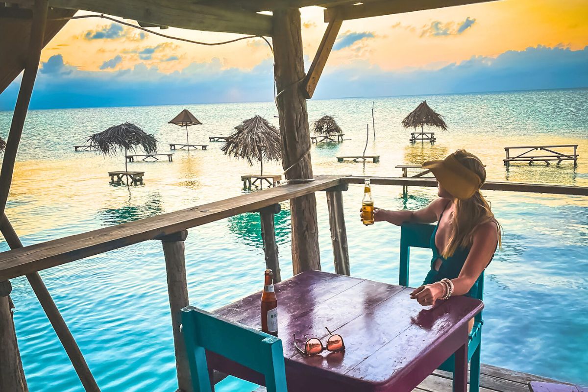 Kate sitting at a wooden table at a waterfront restaurant in Belize, holding a beer, and looking out at the sunset on the horizon. She's Contemplating Belize Travel Tips to share wither her readers.