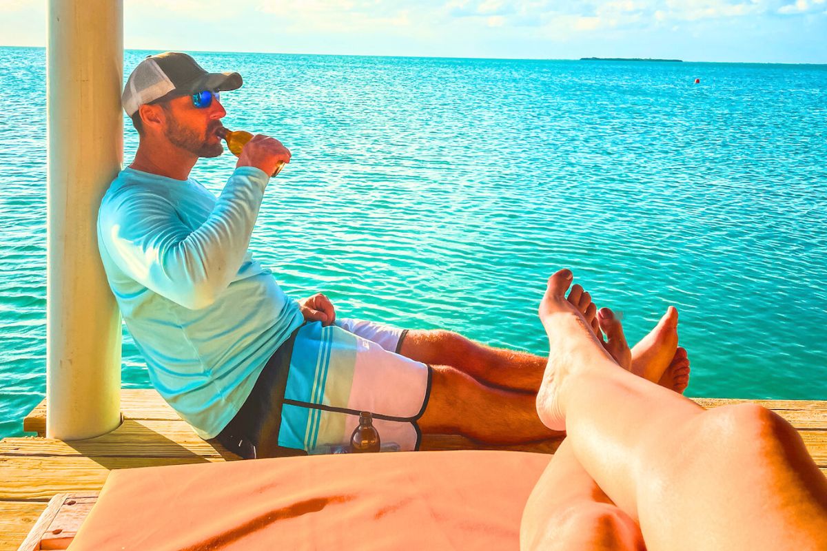 Kate's husband in a ball cap and sunglasses sits on a dock, leaning against a post and drinking a beer, with turquoise water stretching out behind him. In the foreground, Kate's crossed legs are visible, relaxing on the dock in the warm sunlight.