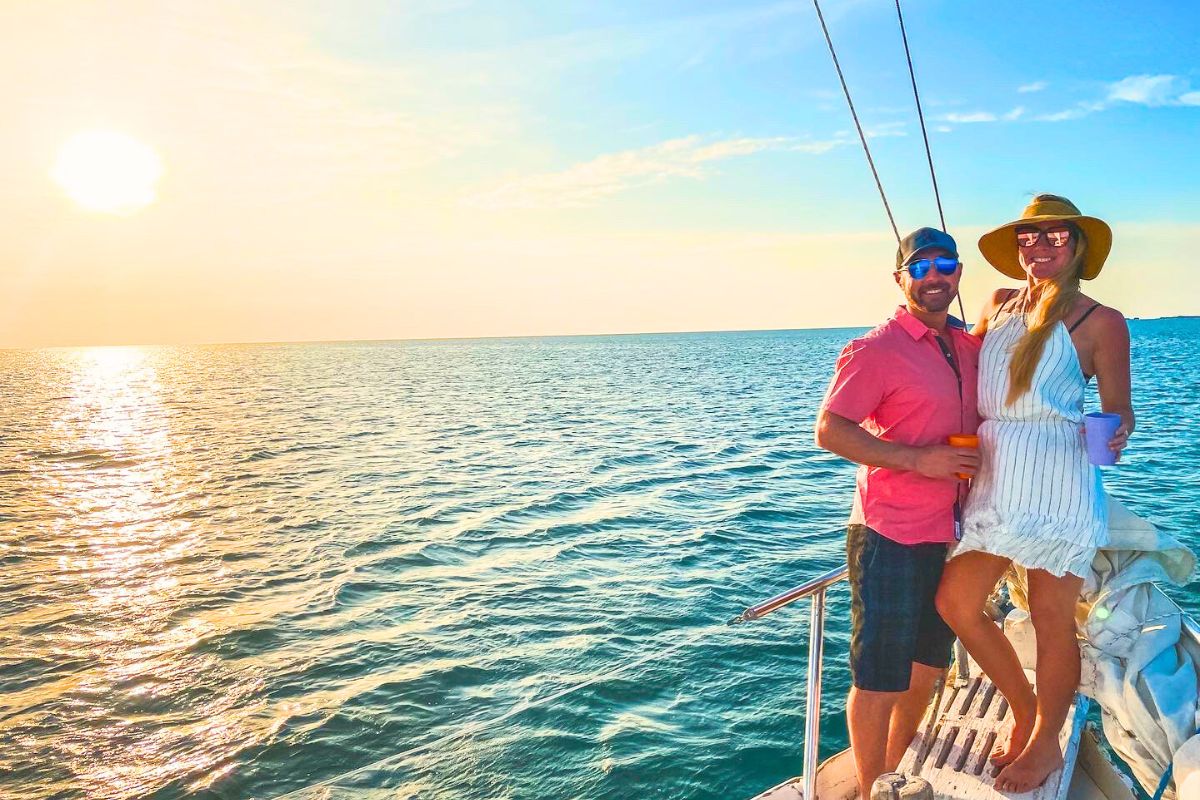 Kate and her husband stand on the deck of a sailboat, holding drinks and enjoying the sunset over the ocean. Kate is wearing a white dress and wide-brimmed hat, while her husband is in a pink shirt and sunglasses. The sun is low on the horizon, casting a warm glow over the water.