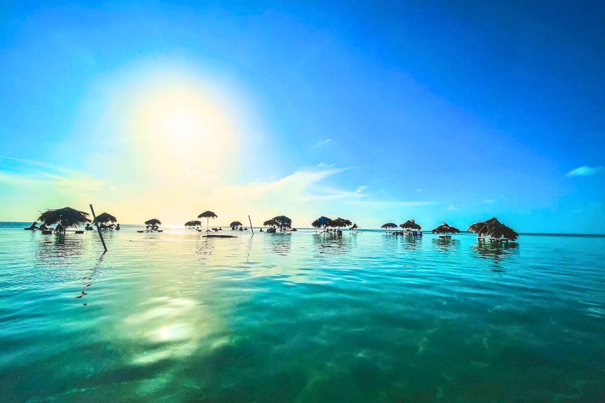 A serene scene of thatched-roof palapas standing over shallow, calm water during sunset on Ambergris Caye Belize. The sun casts a golden glow on the water, creating a peaceful and picturesque setting. The sky is clear with a gradient from blue to warm hues near the horizon.