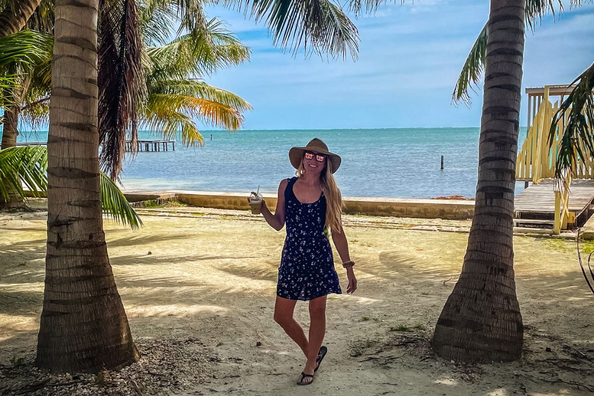 Kate enjoying a sunny day on the beach in Caye Caulker, Belize, wearing a sun hat and holding a drink, with palm trees and the ocean in the background.