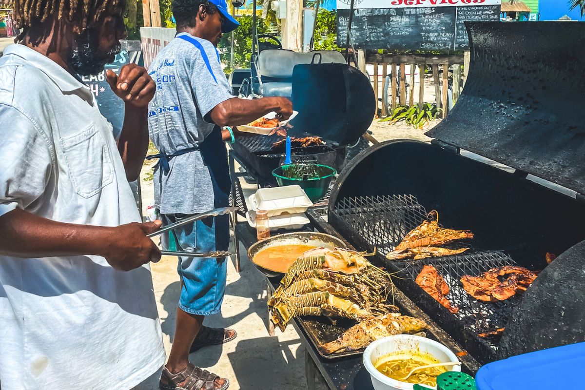 Two men grilling seafood, including lobsters and fish, at a street food stand in Caye Caulker, Belize, with various dishes and sauces being prepared on large barbecue grills.