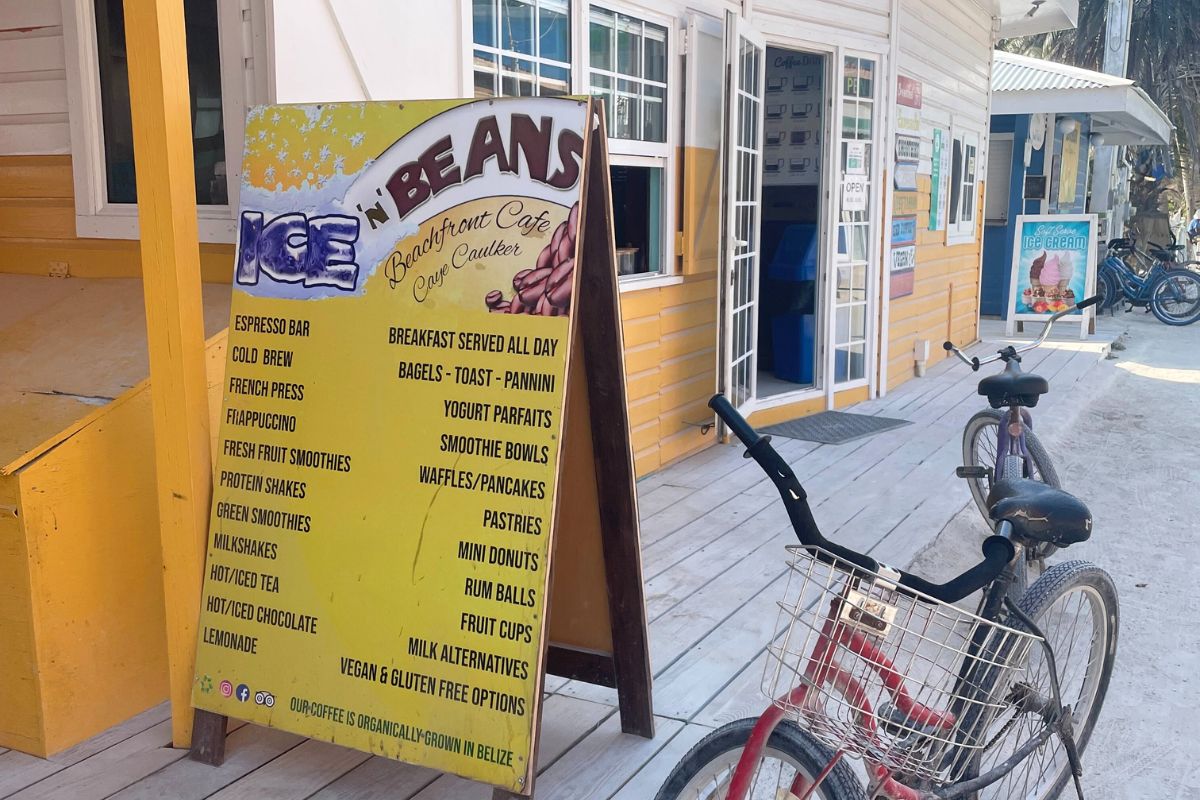 A menu board outside Ice 'n' Beans, a beachfront café in Caye Caulker, Belize, listing various drinks and food items. A bicycle is parked in front of the yellow and white building, adding to the casual, beachy atmosphere.