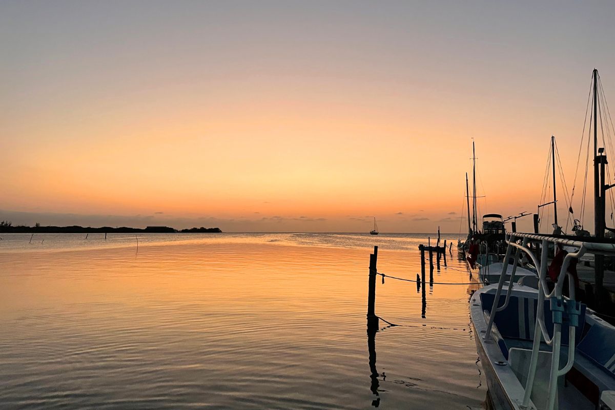 A peaceful sunset over the calm waters of Caye Caulker, Belize, with silhouettes of boats and docks creating a serene and picturesque scene. The sky is painted in soft orange and pink hues, reflecting on the water.