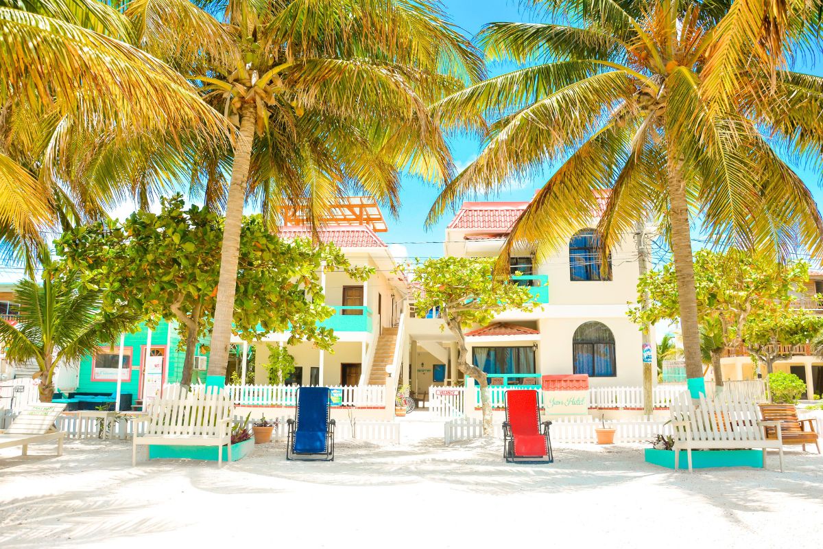 A bright and colorful beachfront hotel called Jan's Hotel, with palm trees providing shade. The sandy courtyard is adorned with white picket fences and lounge chairs in red and blue.