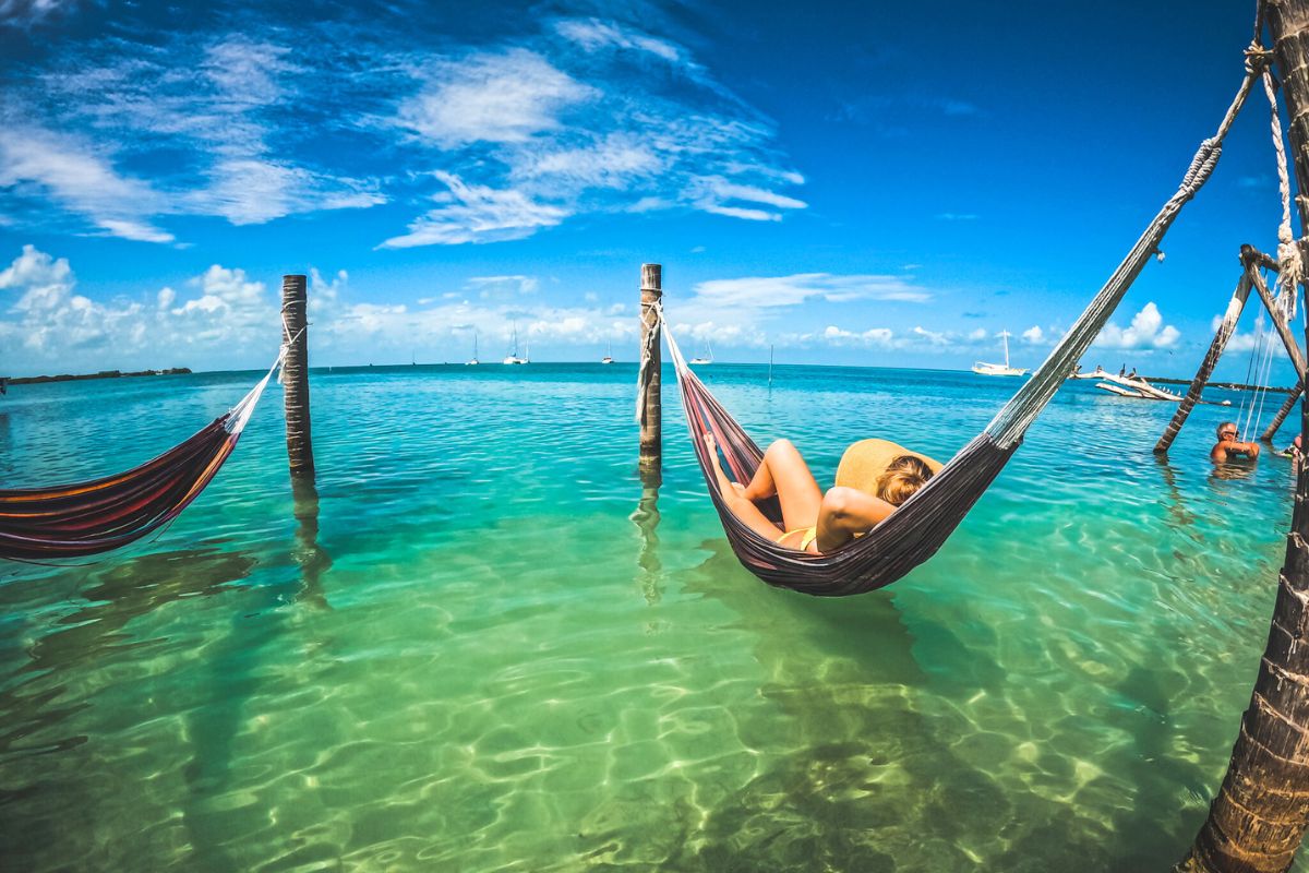 Kate relaxing in a hammock strung between poles over the clear turquoise waters of Caye Caulker, Belize. The scene is set under a bright blue sky with a few sailboats in the distance.