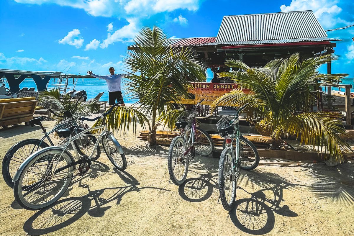 Bicycles parked outside a beachside bar in Caye Caulker, Belize, with a person joyfully spreading their arms in the background. The scene is vibrant with palm trees, blue skies, and the ocean, creating a laid-back, tropical vibe.