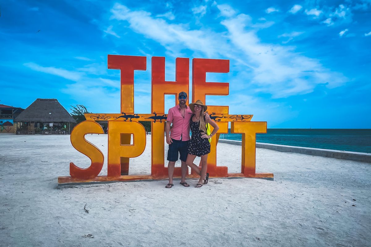 Kate and her husband posing in front of "The Split" sign on the beach in Caye Caulker, Belize, with a clear blue sky and ocean in the background.