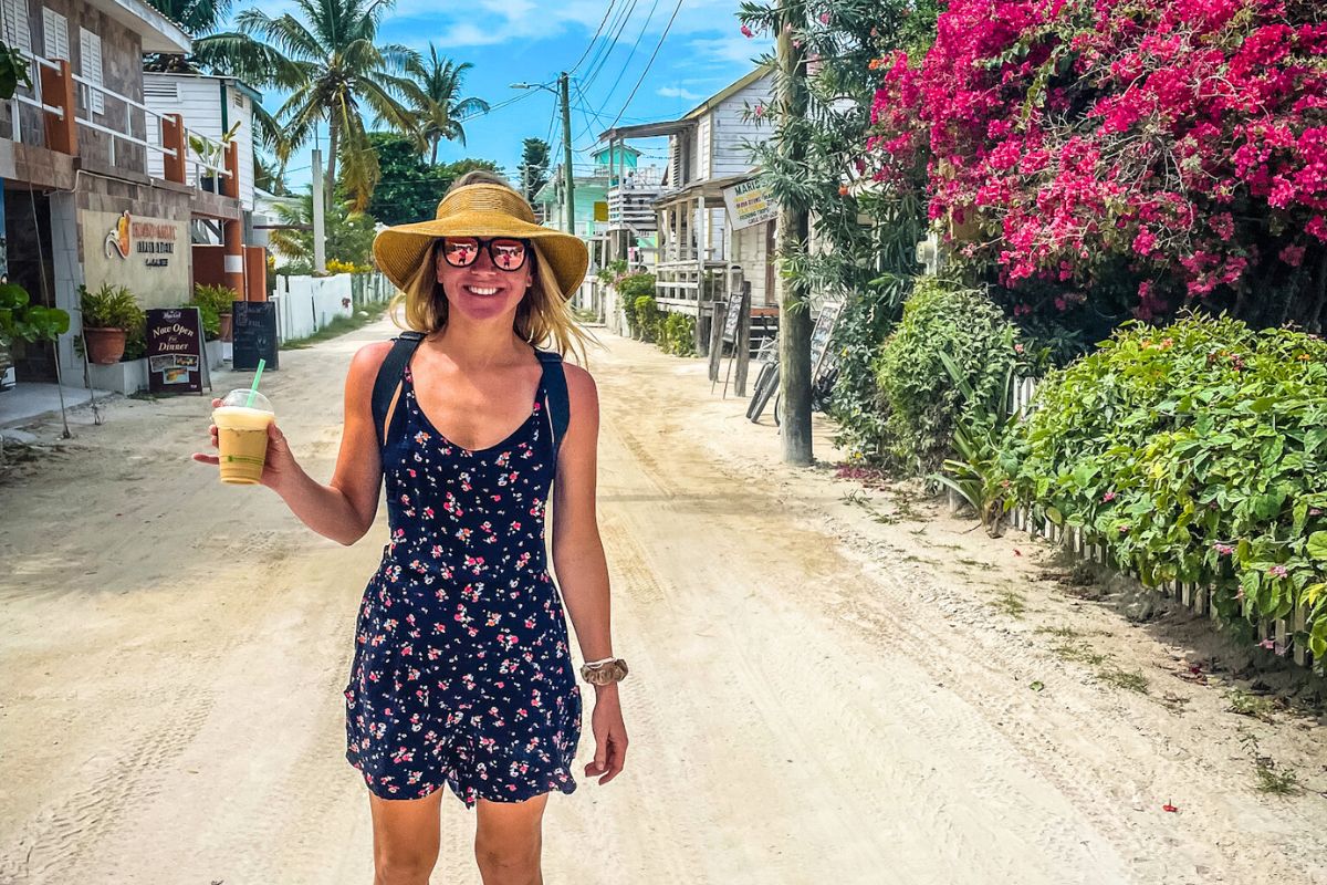 Kate walking down a sandy street in Caye Caulker, Belize, wearing a sun hat and a floral dress. She is smiling and holding a drink, with tropical plants and charming buildings lining the street behind her.