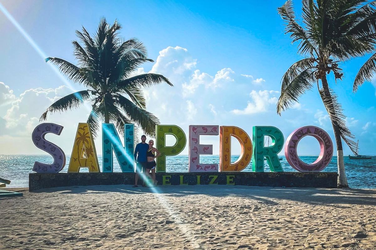 Kate and her husband posing in front of the colorful "San Pedro" sign on the beach in Belize. The backdrop features palm trees, a bright blue sky, and the ocean.