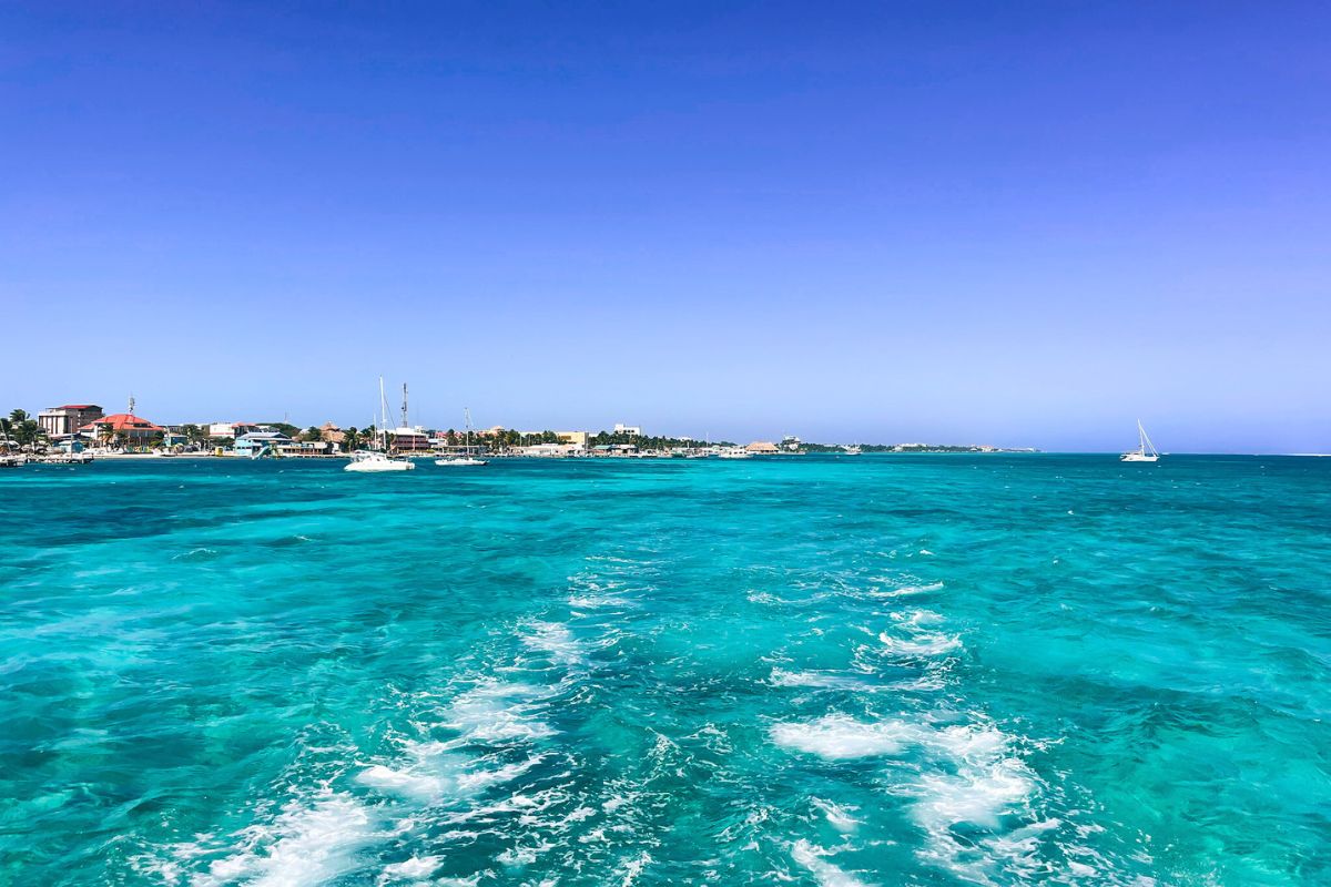 A view of the turquoise waters and the shoreline of San Pedro, Belize, seen from the water. The vibrant blue sea contrasts with the clear sky, and the town's buildings and sailboats are visible in the distance