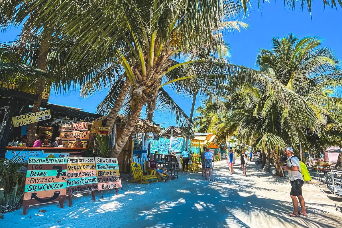 A bustling street scene in Caye Caulker, Belize, lined with palm trees and vibrant local food stalls. Signs advertise various dishes like tacos, pupusas, and stew chicken. People are walking and enjoying the laid-back, tropical atmosphere under the bright blue sky.