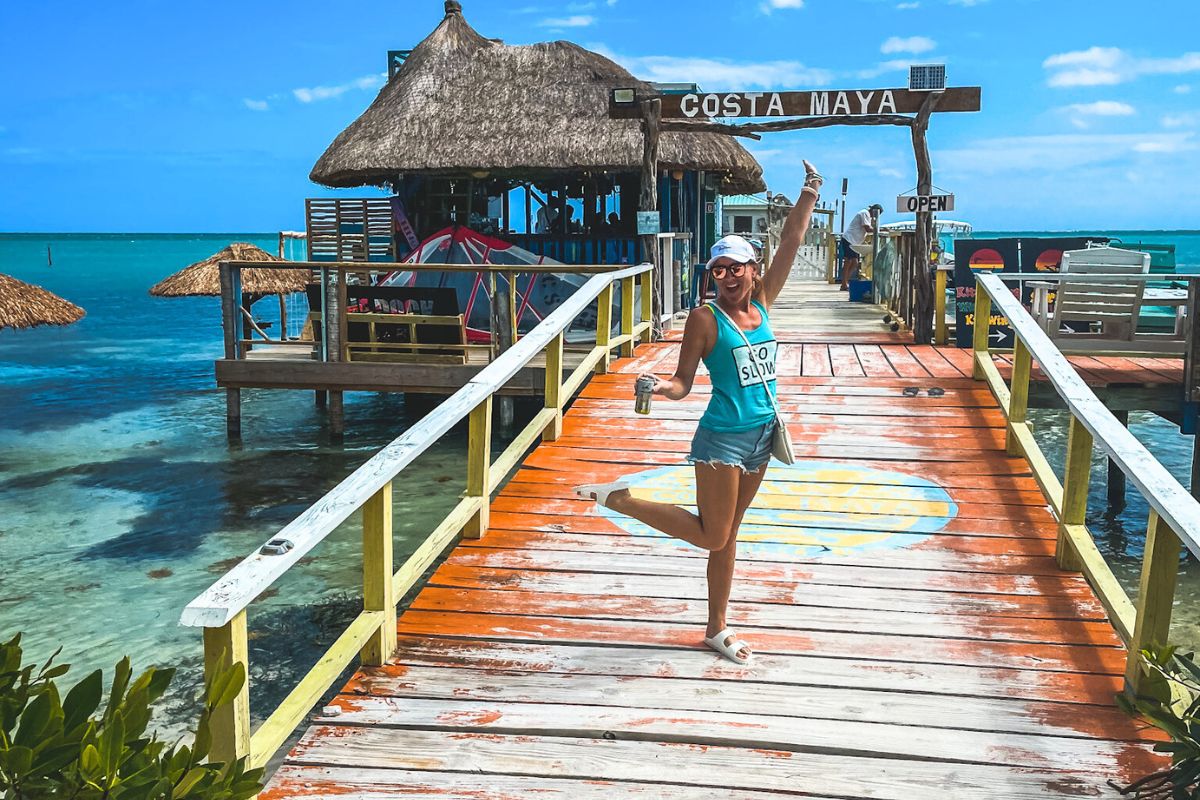 Kate happily posing on a colorful wooden pier leading to Costa Maya Beach Club in Caye Caulker, Belize. She's wearing a turquoise tank top, denim shorts, and a white cap, holding a drink with the beautiful ocean and thatched-roof structures in the background.