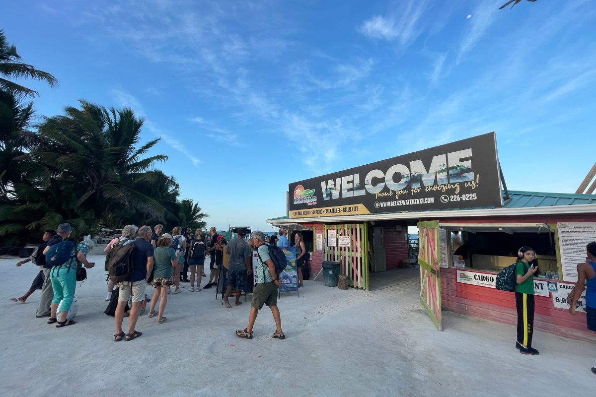 People gathering at the water taxi dock in Caye Caulker, Belize, with a "Welcome" sign and palm trees in the background.