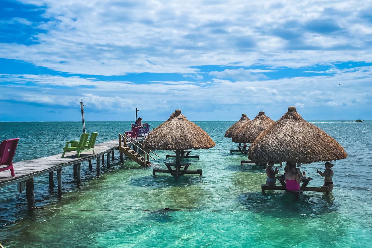 Colorful chairs on a wooden pier and thatched-roof tables in the clear turquoise waters of Caye Caulker, Belize, with people enjoying drinks.