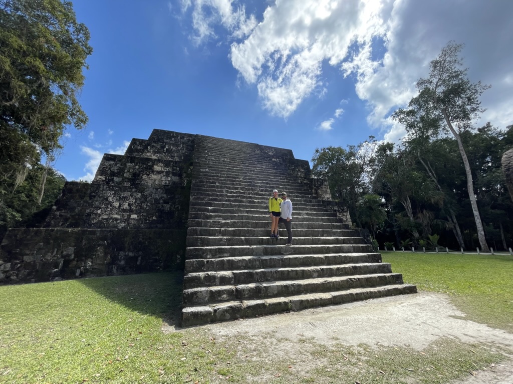 Kate and her husband standing on the steps of a Mayan pyramid surrounded by jungle during a Tikal day trip from Belize. Both are wearing hiking gear they packed for Belize