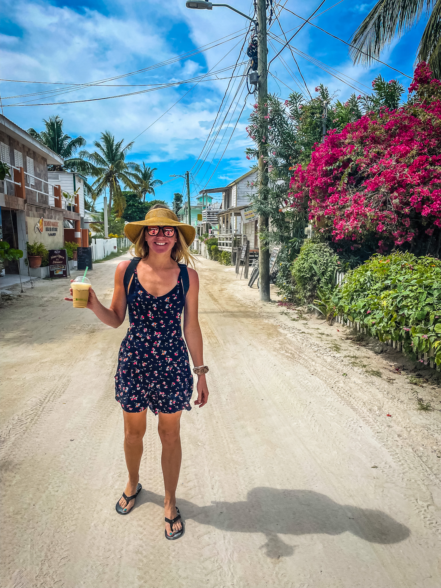 Woman in a floral dress and sun hat holding a drink, walking down a sandy street lined with palm trees and colorful flowers in Belize. Perfect example of a Belize packing list outfit.