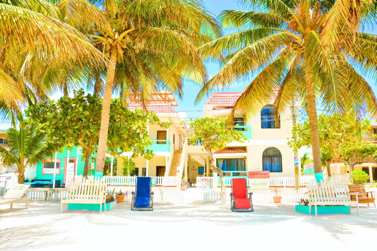 Jan's Hotel in Caye Caulker, Belize, is framed by tall palm trees and lush greenery, with colorful beach chairs and benches placed in the sandy courtyard. The building features vibrant blue and turquoise accents, creating a cheerful and inviting atmosphere under a bright, clear sky.