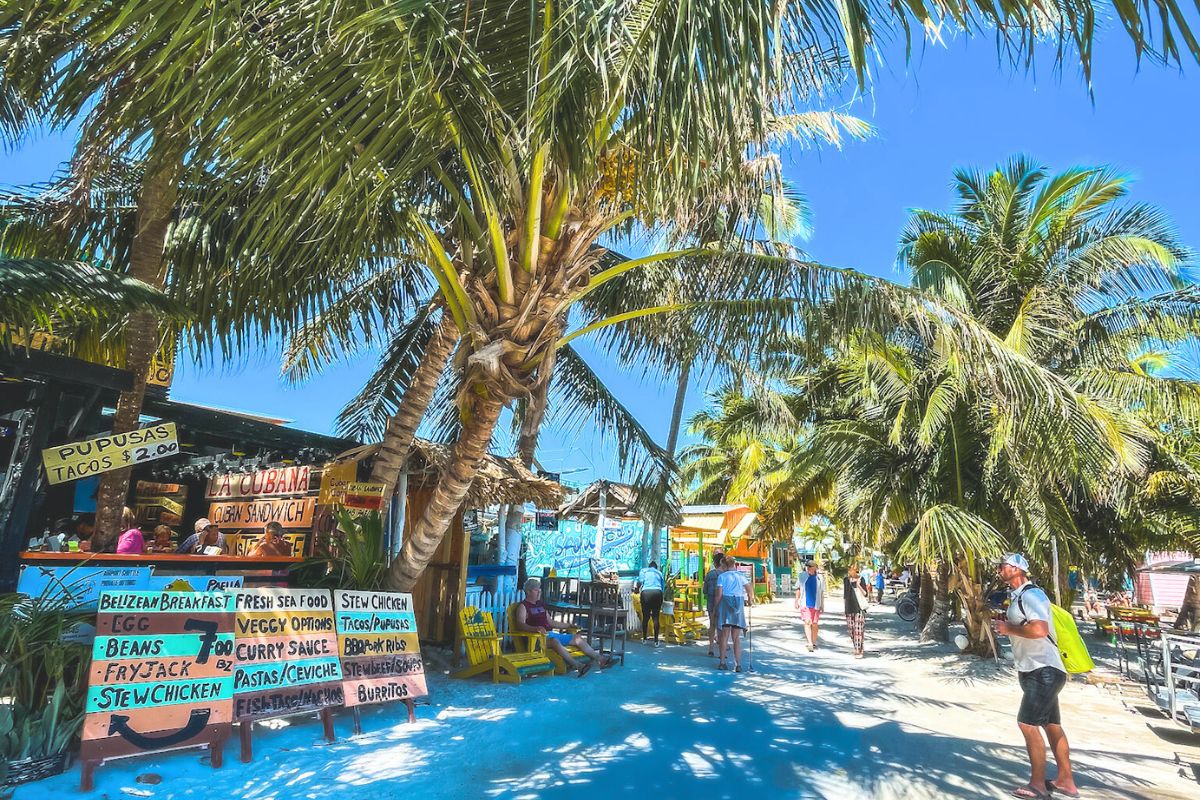 A bustling street scene in Caye Caulker, Belize, shows food stalls under palm trees offering items like pupusas, tacos, sandwiches, and fresh seafood. The vibrant atmosphere includes tourists exploring the area, with colorful signs and beach chairs adding to the lively setting.