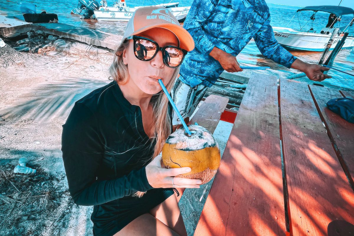 Kate in a baseball cap and sunglasses drinks from a fresh coconut with a straw while sitting at a picnic table near the waterfront in Caye Caulker, Belize. Boats are visible in the background, adding to the tropical, laid-back vibe of the scene