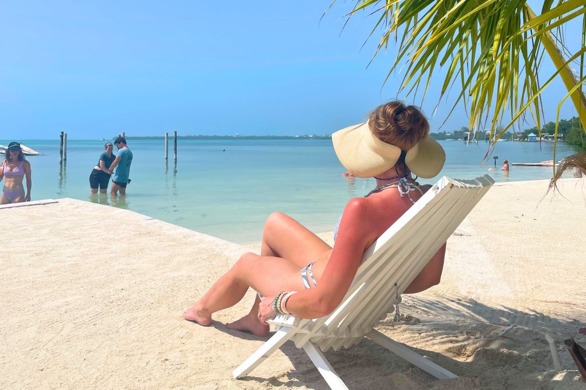 Kate in a wide-brimmed sun hat relaxes on a white beach chair facing the calm, shallow waters in Caye Caulker, Belize. Other beachgoers are seen wading in the water and enjoying the serene, sunny day under clear blue skies.