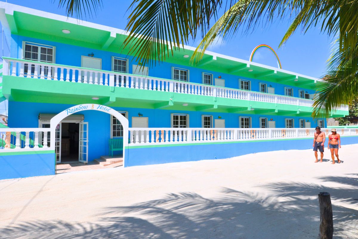 The Rainbow Hotel in Caye Caulker, Belize, features a vibrant blue and green exterior with white balconies and a rainbow arch above the entrance. Palm tree shadows fall on the sandy path in front, where two people in swimwear walk by, enjoying the sunny day.