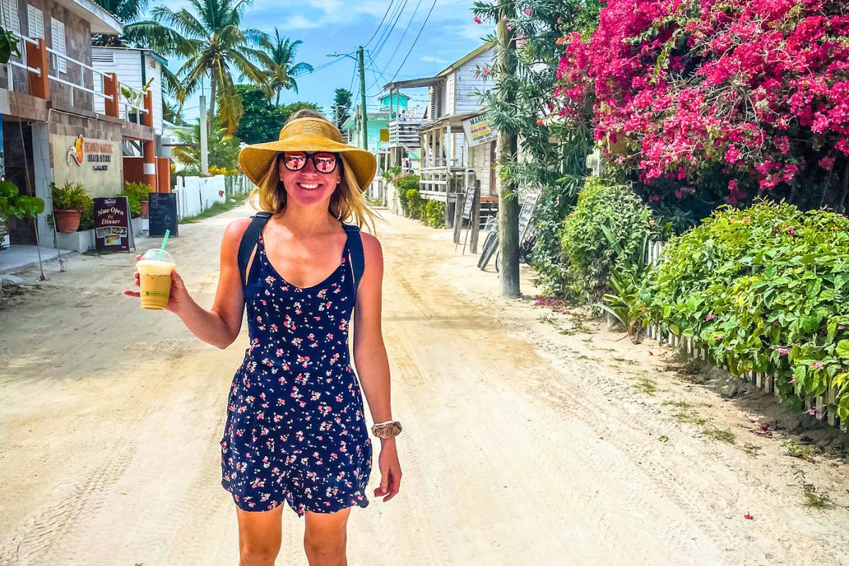 Kate in a floral dress and straw hat smiles while holding a cold drink on a sandy street lined with tropical plants and vibrant pink flowers in Caye Caulker, Belize. The background features rustic buildings and palm trees, creating a relaxed and picturesque setting. Answering the question, "Is Caye Caulker safe?"