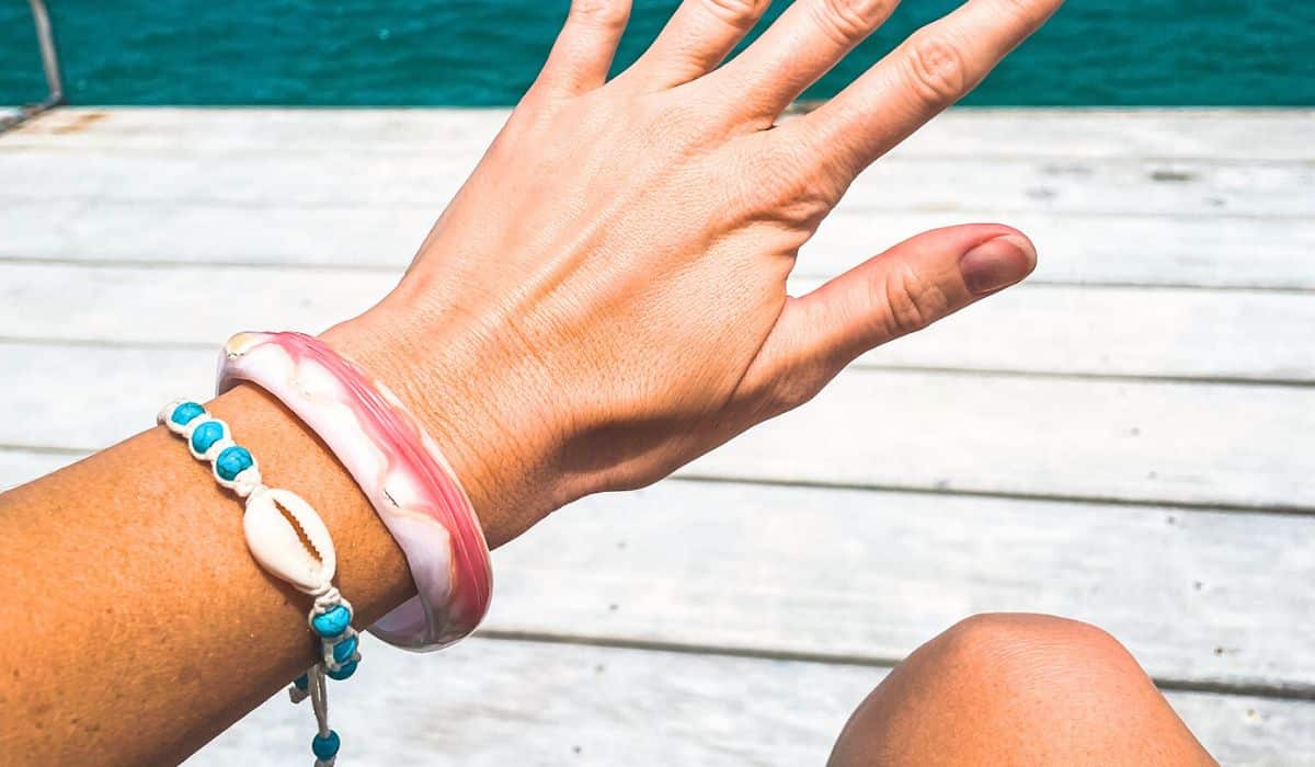 Close-up of Kate's wrist adorned with a pink and white conch shell bracelet and a beaded bracelet with a cowrie shell. She is sitting on a wooden dock with the turquoise ocean in the background, enjoying the tropical scenery.