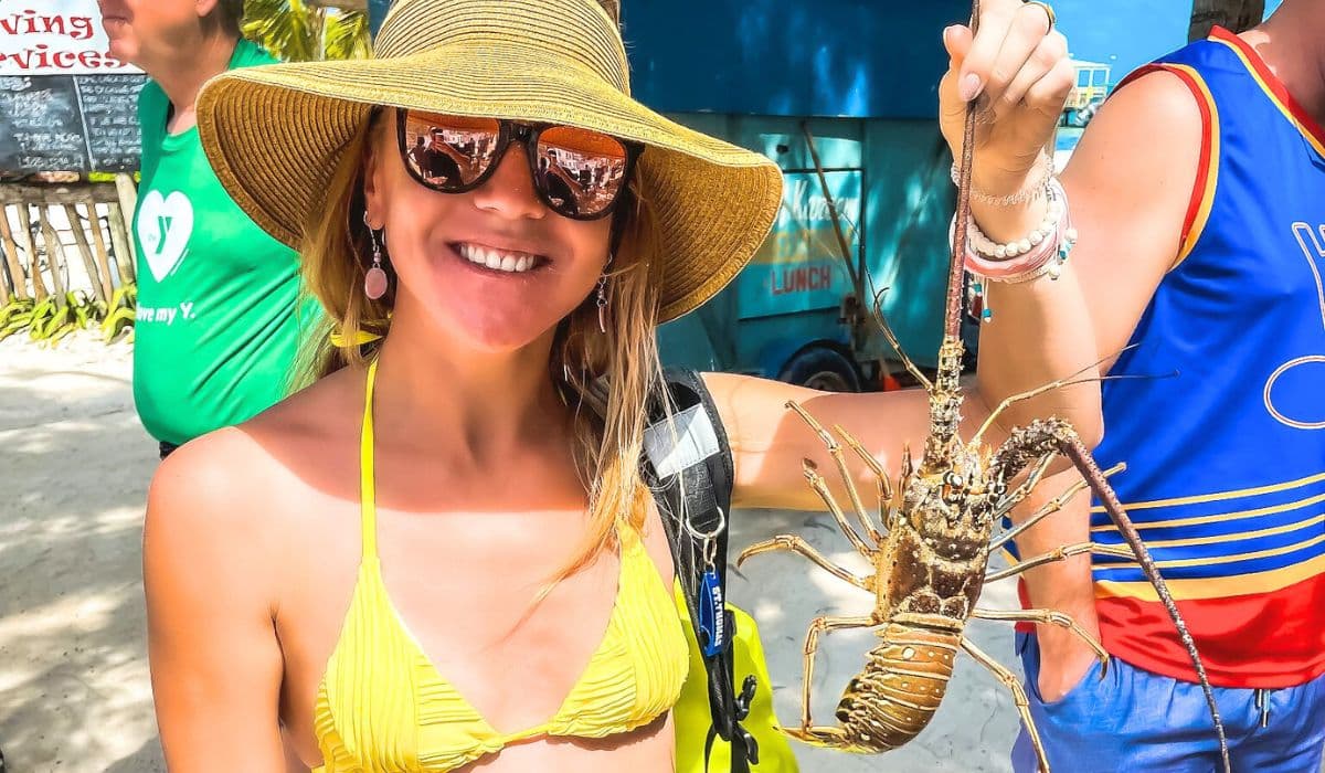 Kate smiling and holding a large lobster at Lobster Fest on Caye Caulker, Belize. She is wearing a yellow bikini top and a sunhat, enjoying the festive atmosphere of the event.