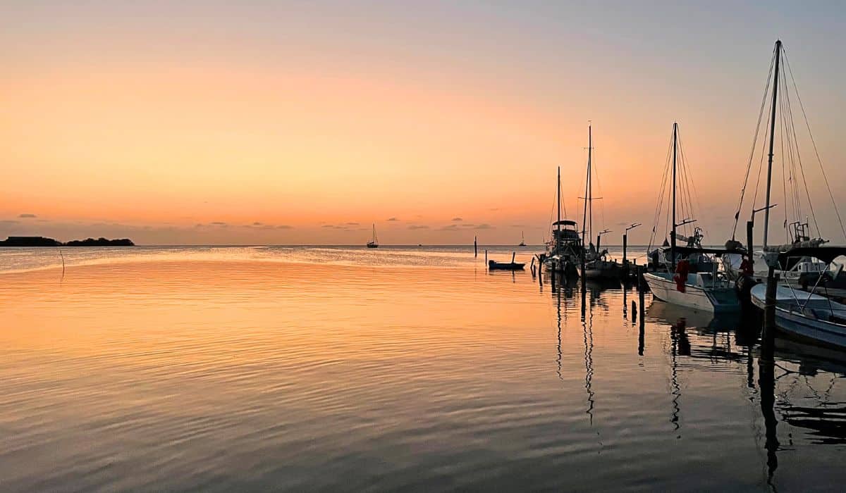 A tranquil sunset view from Caye Caulker, Belize, with a warm orange and pink sky reflecting on the calm waters. Sailboats and small boats are docked at the pier, creating a serene and picturesque scene as the day ends.