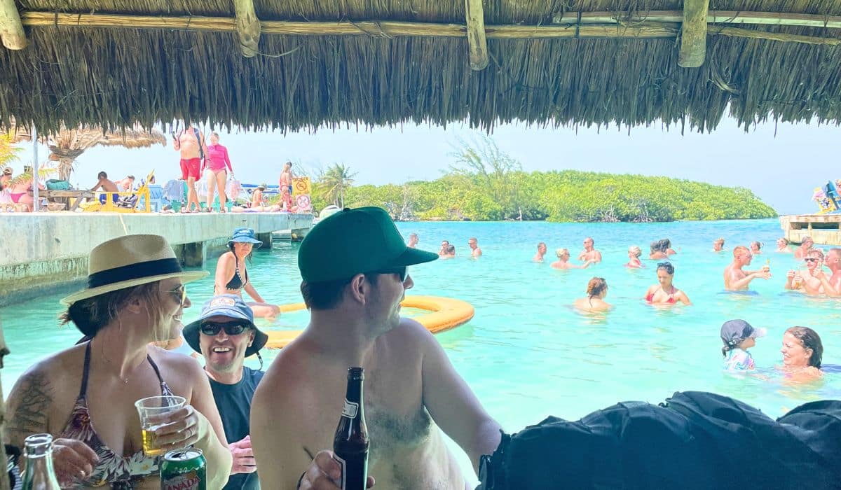 People enjoying drinks and socializing at The Split in Caye Caulker, Belize, a popular spot featuring clear turquoise waters and a lively atmosphere. Patrons relax under a thatched roof bar while others swim and lounge in the shallow water.