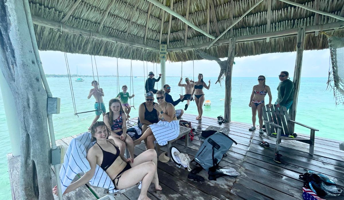 A group of people relaxing and enjoying their time at the Northside Beach Club in Caye Caulker, Belize. They are sitting under a thatched roof structure with swings hanging over the water, creating a fun and laid-back atmosphere.