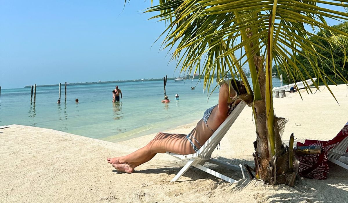 Kate relaxing on a lounge chair under a palm tree on a sandy beach in Caye Caulker, Belize. She is enjoying the shade and the serene view of the calm, shallow waters where other people are swimming and wading.