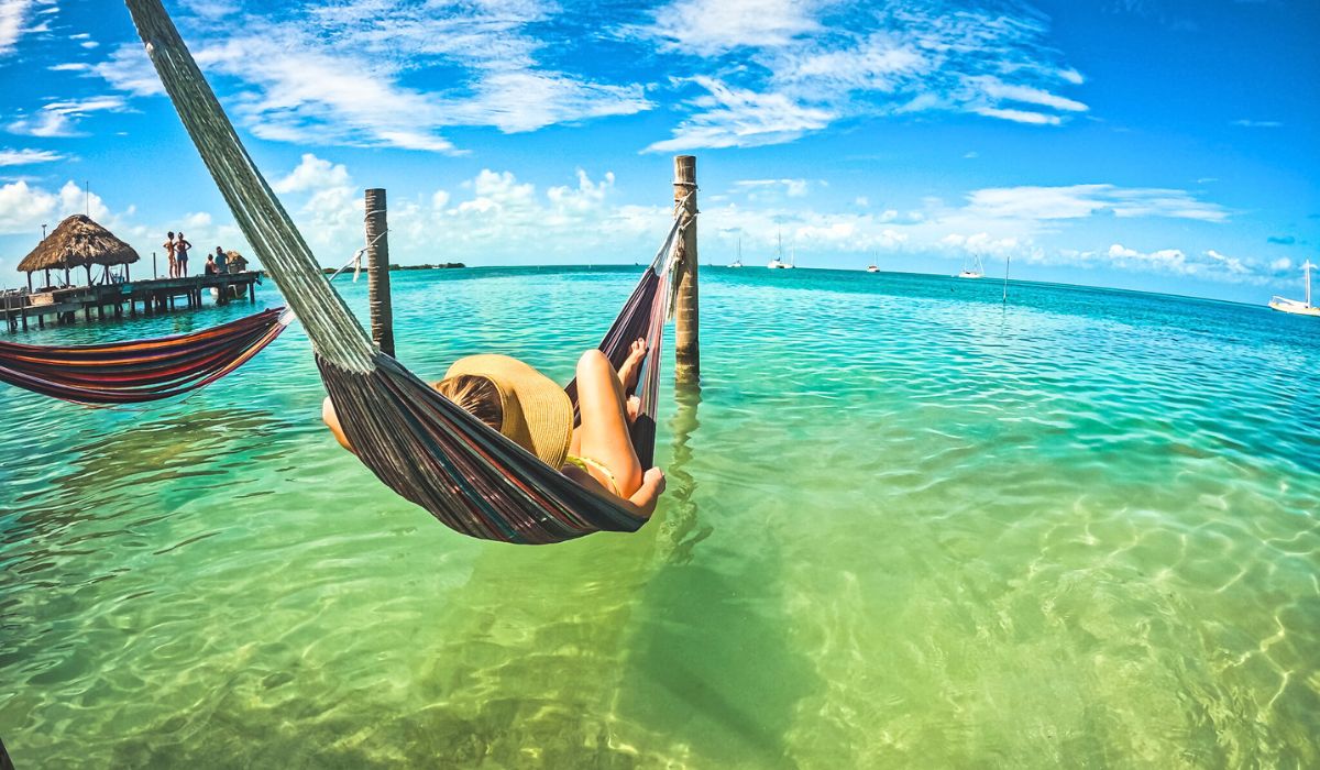 Kate relaxing in a hammock suspended over clear, turquoise waters in Caye Caulker, Belize. They are wearing a sunhat and enjoying the serene view of the ocean with a distant dock and boats under a bright blue sky.