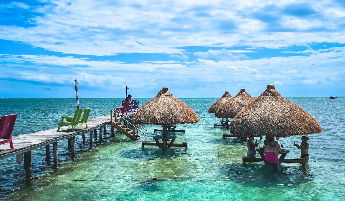 People enjoying drinks at the Sip N Dip Bar in Caye Caulker, Belize. The bar features thatched-roof huts with tables in the shallow, clear water, and colorful chairs on a wooden pier extending into the ocean. The scene is set against a bright blue sky and turquoise sea