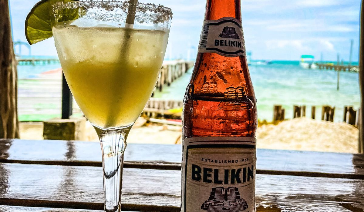 A refreshing margarita with a lime wedge and a bottle of Belikin beer on a wooden table, overlooking a beach and the ocean in Caye Caulker, Belize. The background features a sandy shore, wooden dock, and the clear blue water under a bright sky.